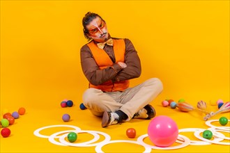 Juggler in waistcoat and with painted face sitting with the juggling objects on a yellow background