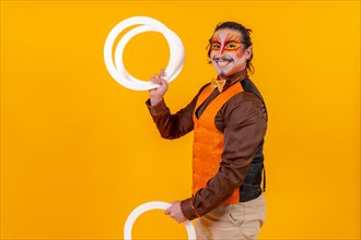 Juggler in a vest and with a painted face juggling hoops on a yellow background