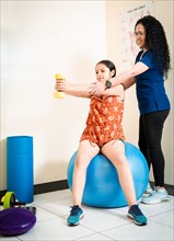 Physiotherapist with female patient holding hand dumbbell sitting on rehab ball. Physiotherapist helping female patient with hand weight sitting on fitness ball