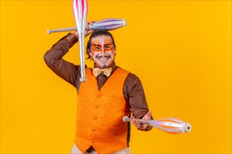 Juggler man in make up vest juggling with maces on a yellow background