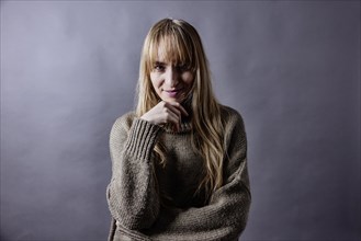 Woman with long blond hair and woollen jumper holds a hand to her chin and smiles at camera