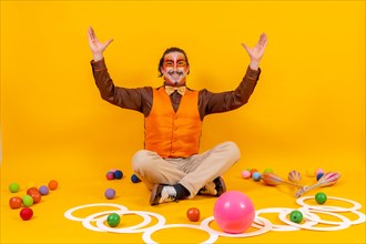 Juggler in a vest and with his face painted sitting with the juggling objects on a yellow background