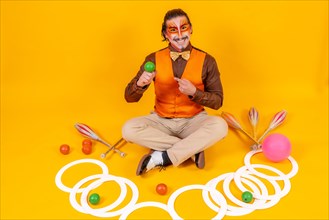 Portrait of a juggler in a vest and makeup sitting with the objects on a yellow background