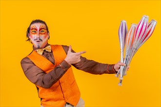 Portrait of a happy juggler man in make up vest juggling with maces on a yellow background
