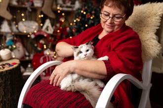 Grandmother is sitting on a rocking chair with a cat against the backdrop of Christmas arrangement. In studio