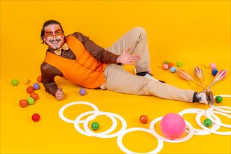 Juggler in a vest and with a painted face lying with the juggling objects on a yellow background