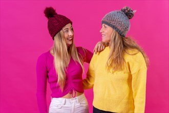 Two blonde caucasian women in wool hats on a pink background looking at each other smiling