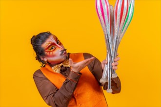 Kiss and portrait of happy juggler man in makeup vest juggling clubs on yellow background