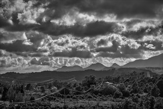 Dramatic sky over rugged mountain silhouettes and woods crossed by power lines in black and white in Spain