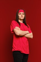 A young adult man in red shirt and head band stands in a studio with arms crossed and looks confidently at the camera against red background