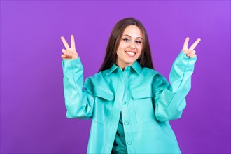 Young woman isolated on purple background smiling and showing victory sign wearing blue windbreaker