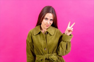 Young woman over pink wall smiling and showing victory sign wearing green windbreaker