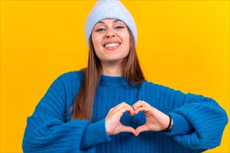 Young brunette woman wearing blue sweater over isolated yellow background smiling in love making heart symbol shape with hands. romantic concept