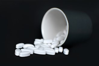 Many white medical pills spilling from a black bottle container on dark black background