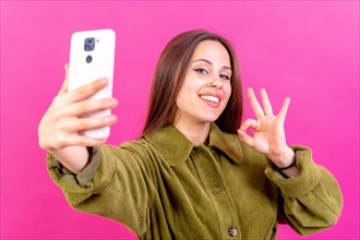 Isolated shot of a caucasian woman using a phone camera taking a selfie doing the ok gesture