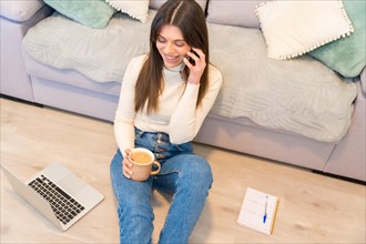 Portrait of woman with a computer sitting on a sofa with a hot coffee talking on the phone