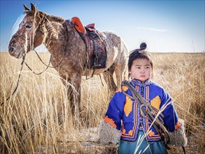 A boy in traditional dress stands next to a horse. Dornod Province