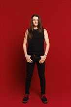 Full body indoor portrait of positive young man with long hair standing against red background