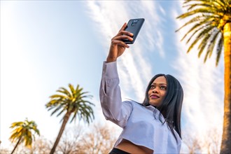 Young african woman posing in white clothes in a tropical place with palm trees at sunset enjoying the summer taking a selfie