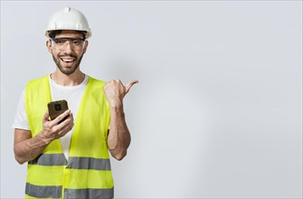 Smiling engineer holding phone and pointing to the side isolated. Young construction engineer holding phone and pointing at blank space. Civil engineer holding cellphone and pointing a promo