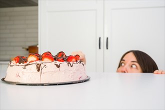 Young woman hiding behind the table eating strawberry shortcake in her home kitchen
