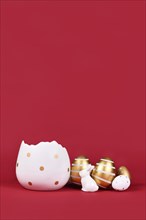Golden and white easter eggs painted with stripes, bunny and egg cup at bottom of dark red background with copy space