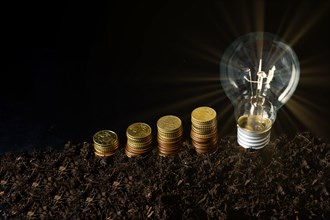 Rising piles of coins with lighted light bulb on black background, concept of rising electricity prices