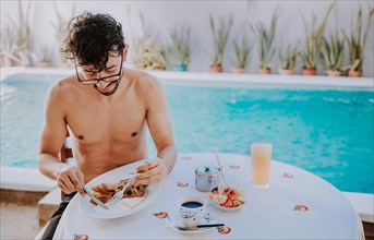 Young man on vacation in hotel having breakfast near swimming pool. Breakfast near the swimming pool. Person having breakfast in the hotel with pool in the background