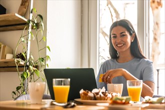 Woman shopping online with computer while having breakfast in the morning by the window with orange juice