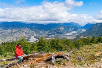 Hiker taking a break on a tree trunk, ascent to the lagoon on Cerro Castillo mountain, the river valley of the Rio Ibanez in the background, Cerro Castillo National Park, Aysen, Patagonia, Chile, Sout...