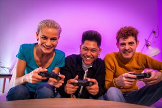 Group of young friends playing video games together on the sofa at home, competing looking at camera