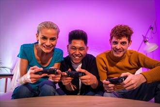 Group of young friends playing video games together on the sofa at home, purple led, competing looking at camera