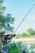 Fisherman with beard and cap on the bank of the river with rod in hand pulling out a carp