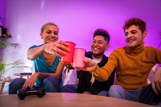 Group of young friends playing video games together on the sofa at home, toasting with colored glasses, young millennials