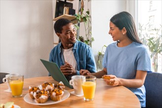 Multi-ethnic couple making purchase online with the computer while having breakfast, next to the window