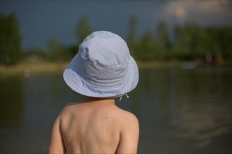 A child standing with his back in a hat by the water and looking into the distance. A four-year-old boy