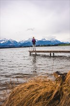 Hiker woman with backpack standing on footbridge at the lake looking at the mountain panorama