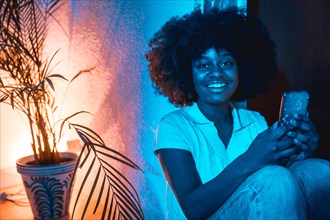 African american woman with afro hair sitting at home on the floor looking at the phone at night with a blue light