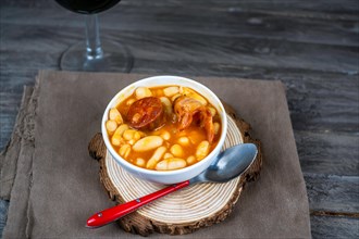 White beans stewed with chorizo in white ceramic bowl on wooden table with a glass of red wine