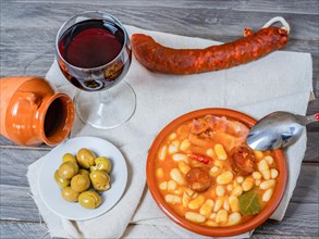 White beans stewed with bacon and chorizo in an earthenware casserole on a wooden table with all the ingredients of the recipe