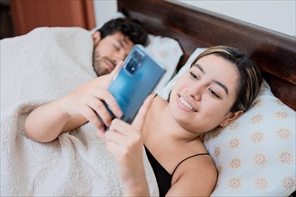 Unfaithful wife with phone while her husband sleeps. Unfaithful woman with phone while the man sleeps. Unfaithful girlfriend with phone while boyfriend sleeps