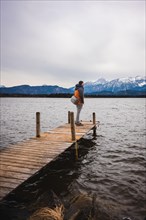 Man standing on footbridge at lake looking at mountain panorama