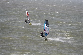 Two recreational windsurfers in black wetsuits practising classic windsurfing along the North Sea coast in windy weather during winter storm