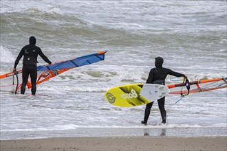 Two windsurfers in black wetsuits entering the water to practise classic windsurfing along the North Sea coast in windy weather during winter storm