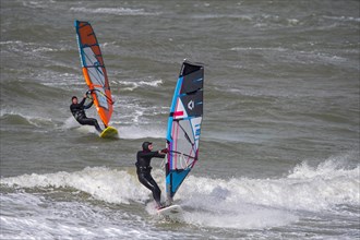 Two recreational windsurfers in black wetsuits practising classic windsurfing along the North Sea coast in windy weather during winter storm