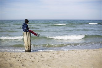 Symbolic photo on the theme of loneliness in childhood. A child sits alone on a wooden post by the sea. Arenshoop