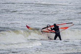 Windsurfer in black wetsuit entering the water to practise classic windsurfing along the North Sea coast in windy weather during winter storm