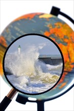 Waves crashing into jetty during storm at sea seen through magnifying glass held against illuminated terrestrial globe