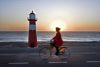 Woman riding bicycle along red lighthouse at sunset