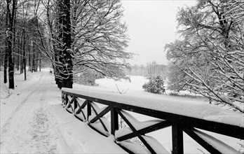 Winter landscape with hiking trail and a wooden bridge railing. Two unrecognisable people are visible in the background. The black and white shot fits the cold mood in the forest